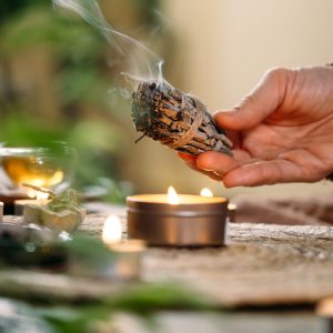Woman hands burning white sage, before ritual on the table with candles and green plants. Smoke of smudging treats pain and stress, clear negative energy and meditation